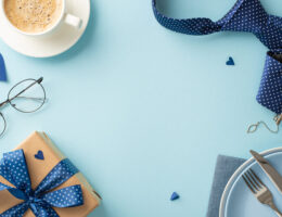 Tischdekoration zum Vatertag mit einer flachen Draufsicht auf Kaffee, Geschirr, Besteck, Krawatte, Herzen, Accessoires, Brillen, Geschenkbox auf einem pastellblauen Hintergrund und einem leeren Rahmen für Text