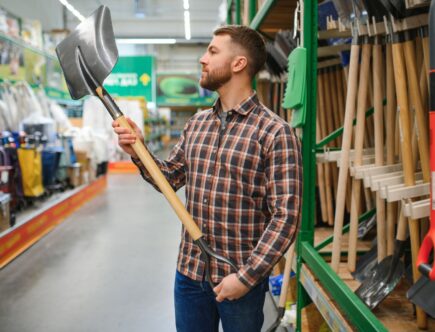 Porträt eines glücklichen jungen Mannes und eines Kunden, der beim Einkaufen im Baumarkt eine Schaufel kauft