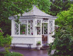 Weißer Gartenpavillon mit Fenstern und dekorativen Pflanzen, umgeben von grünen Bäumen – pavillon kaufen