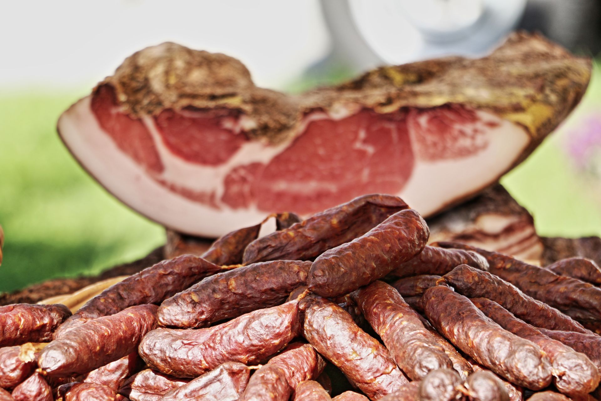 Geräucherte Kaminwurzen vor einem Stück Speck – traditionelle Spezialitäten aus Südtirol, perfekt zum Online-Kauf.