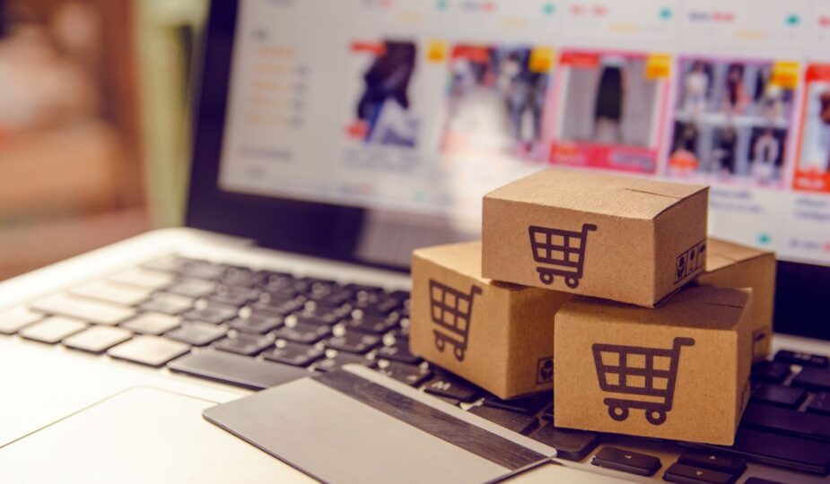 Symbolbild fuer Damenmode online kaufen mit Versandkartons auf Laptop und geoeffnetem Onlineshop im Hintergrund